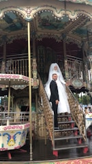 A bride in a white gown and a groom in a black suit are standing on the steps of a vintage carousel. The carousel features ornate decorations with intricate patterns and pastel colors.