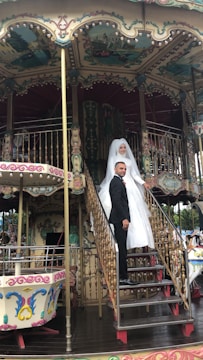 A bride in a white gown and a groom in a black suit are standing on the steps of a vintage carousel. The carousel features ornate decorations with intricate patterns and pastel colors.