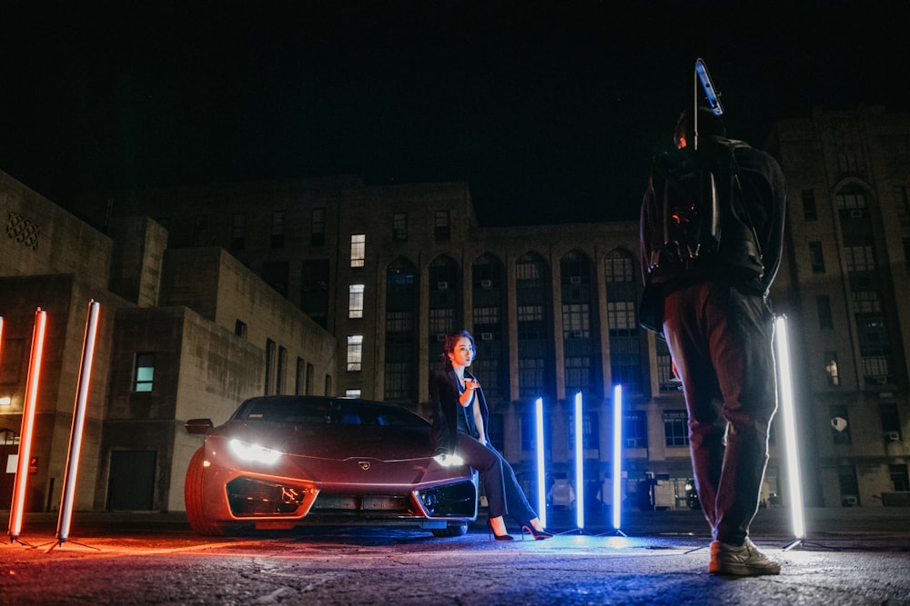 man in black leather jacket and black pants standing beside red car during night time