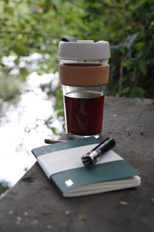 A reusable coffee cup with a cork sleeve and white lid stands on a flat surface outdoors. The cup contains a dark liquid, likely coffee. Beside it lies an open notebook with a green cover and a pen resting on top. The background is filled with lush green foliage, suggesting a garden or park setting.