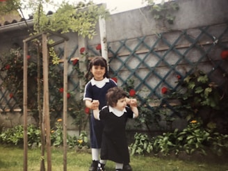 Two children, one older and one younger, are standing in a garden. The older child is wearing a blue dress with a white collar and is holding the younger child, who is dressed in black and white. They are surrounded by greenery and red flowers on a trellis, with a wooden structure to their left.