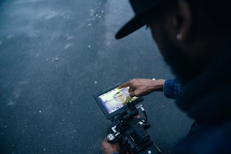 A person pointing at a touchscreen device mounted on a camera setup, seemingly mid-operation or adjustment. The background is a dark, textured surface adding contrast to the scene.