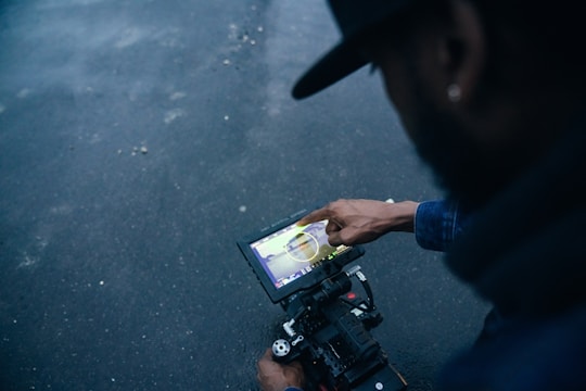 A person pointing at a touchscreen device mounted on a camera setup, seemingly mid-operation or adjustment. The background is a dark, textured surface adding contrast to the scene.