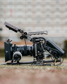 A professional video camera setup is positioned on the ground, equipped with a large lens, a matte box, and various rigging components. The camera has cables attached and is mounted on a stabilizing frame. The background is blurred, suggesting an outdoor setting with soft, neutral colors.