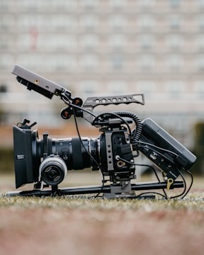 A professional video camera setup is positioned on the ground, equipped with a large lens, a matte box, and various rigging components. The camera has cables attached and is mounted on a stabilizing frame. The background is blurred, suggesting an outdoor setting with soft, neutral colors.