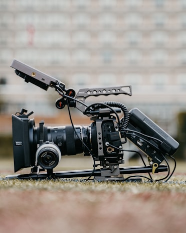 A professional video camera setup is positioned on the ground, equipped with a large lens, a matte box, and various rigging components. The camera has cables attached and is mounted on a stabilizing frame. The background is blurred, suggesting an outdoor setting with soft, neutral colors.