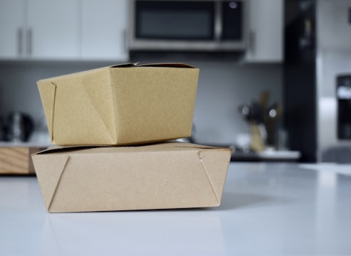 Two brown cardboard takeout boxes stacked on a white countertop in a modern kitchen. The background features blurred elements like a microwave, cabinets, and utensils.