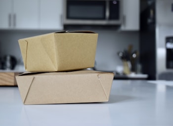 Two brown cardboard takeout boxes stacked on a white countertop in a modern kitchen. The background features blurred elements like a microwave, cabinets, and utensils.