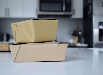 Two brown cardboard takeout boxes stacked on a white countertop in a modern kitchen. The background features blurred elements like a microwave, cabinets, and utensils.