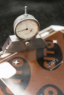 A close-up of a dial gauge measuring tool set at a precise angle on a metal surface, likely used for industrial or engineering purposes. The background includes a metallic board with printed numbers and symbols.