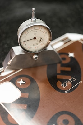 A close-up of a dial gauge measuring tool set at a precise angle on a metal surface, likely used for industrial or engineering purposes. The background includes a metallic board with printed numbers and symbols.