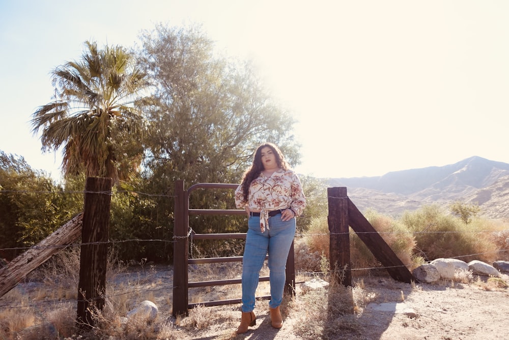 woman in gray sweater and blue denim jeans standing beside brown wooden fence during daytime