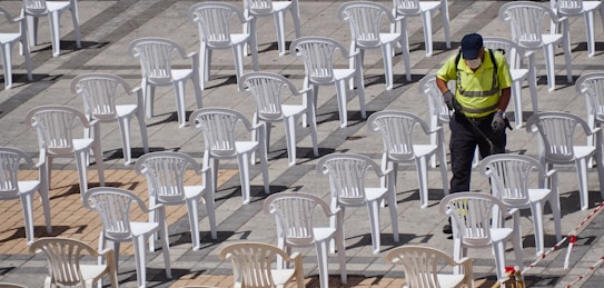 Rows of white plastic chairs are arranged on a checkered pavement with a person in a fluorescent yellow vest and black pants working amidst them, wearing gloves and a mask.