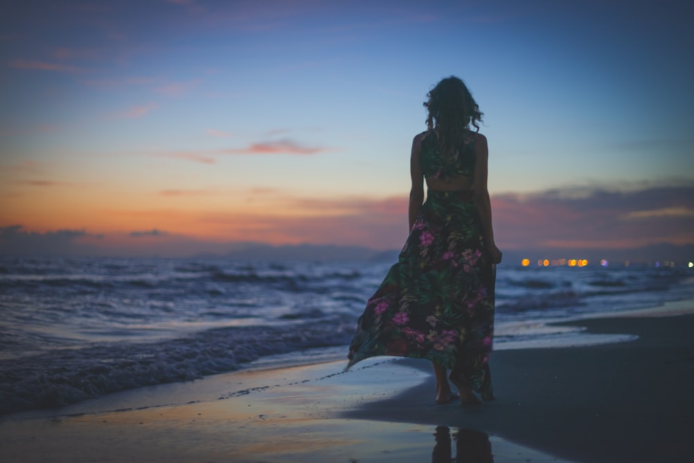 femme en robe à fleurs noire et rouge debout sur la plage pendant le coucher du soleil