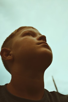 A close-up of a young person looking upwards against a light blue sky background, creating a contemplative and hopeful atmosphere.