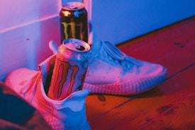 A pair of white sneakers with textured soles rest on a wooden floor. Inside one of the sneakers, there is a can of energy drink with a colorful design. A second can is placed beside the shoes, partially visible. The image is lit with a vibrant pinkish-blue lighting, creating a bold contrast.