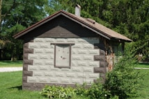 A small, rustic shed with a decorative design on the siding, surrounded by lush greenery. The roof is pitched, and there is a single window with an illustration of a duck and birds. Ivy climbs up one side of the structure, adding to the natural setting.