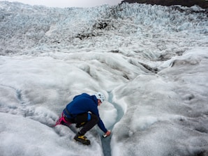 A person is kneeling on a vast icy glacier, using a small tool to explore a crevasse. The surface is rugged and textured with a mix of ice and snow. The person is dressed in outdoor gear with a helmet for safety.