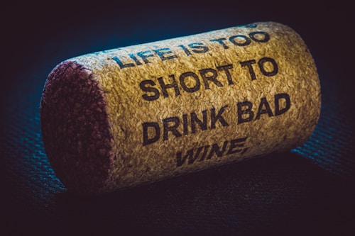 A close-up view of a wine cork with the text 'LIFE IS TOO SHORT TO DRINK BAD WINE' embossed on it. The cork rests on a textured dark surface, and is illuminated by subtle lighting, highlighting its rough texture.