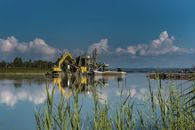 A serene water body reflects a large, yellow excavation machine and industrial structure set against a backdrop of lush trees and a clear blue sky with fluffy clouds. Tall grass lines the foreground, adding a natural element to the otherwise industrial scene.