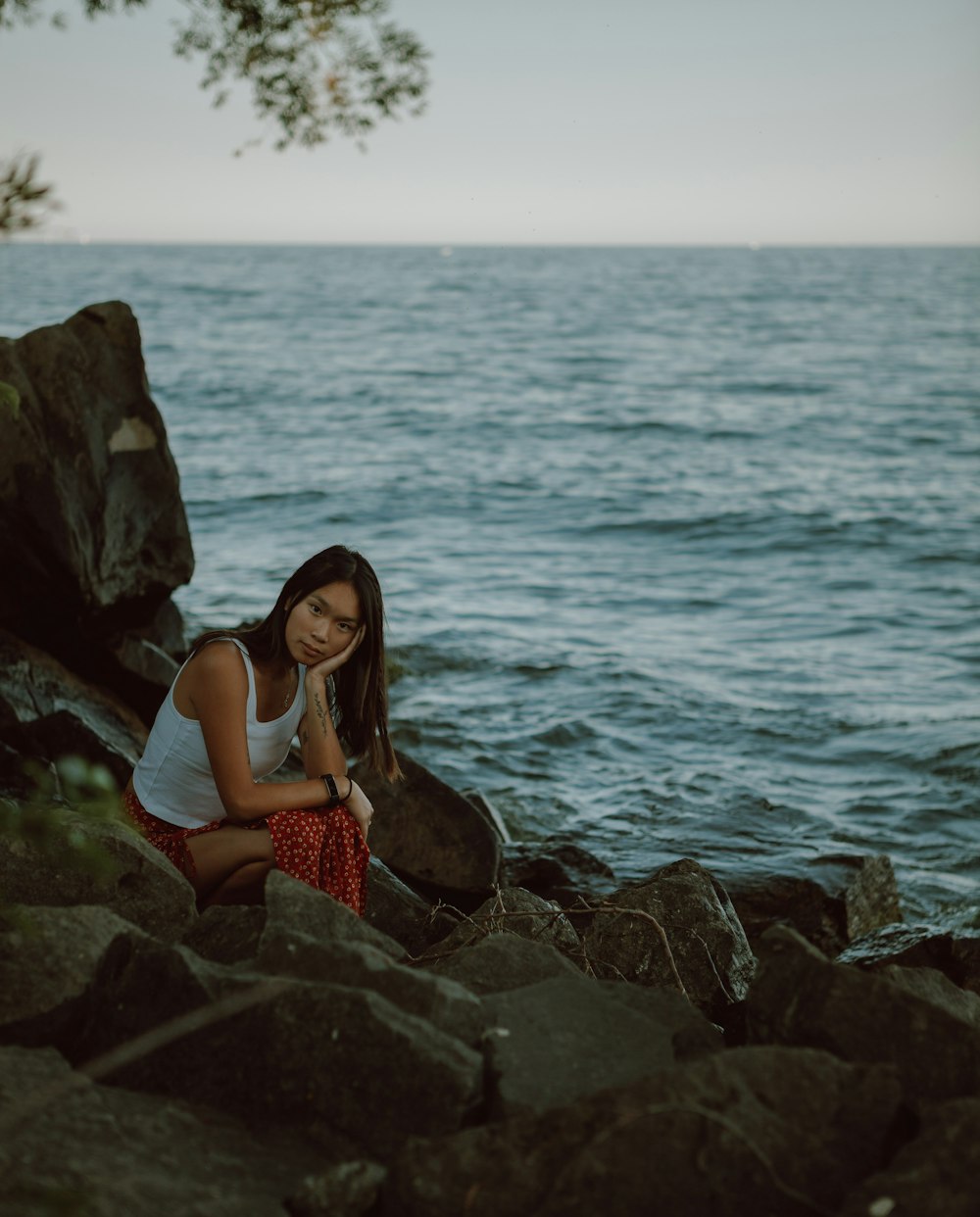 白いタンクトップと赤いスカートを着た女性が昼間、海辺の岩の上に座っている