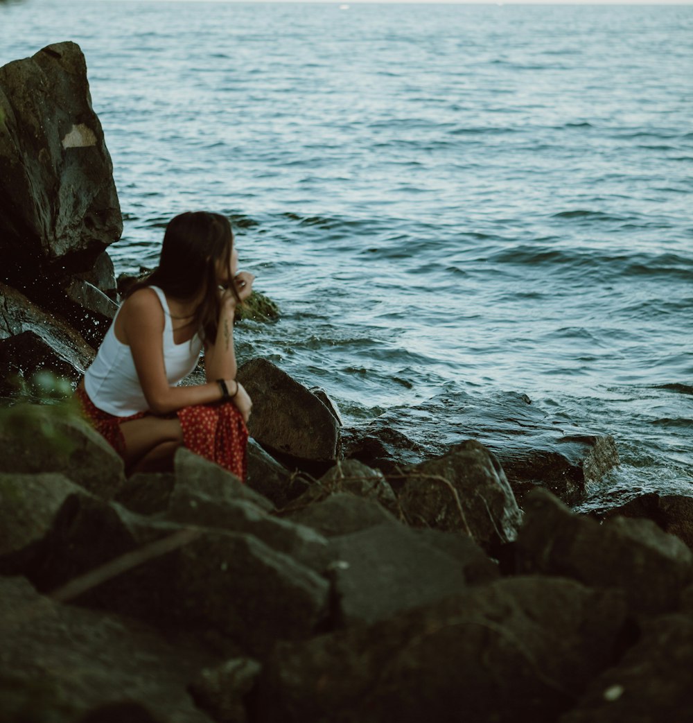 woman in white tank top and red skirt sitting on rock by the sea during daytime