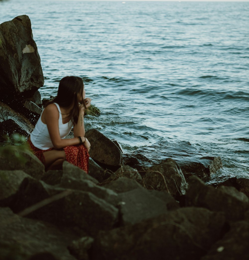 femme en débardeur blanc et jupe rouge assise sur le rocher au bord de la mer pendant la journée