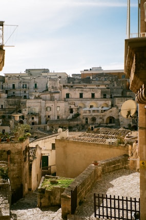 A historic townscape featuring stone buildings with rustic facades, characterized by their aged, weathered appearance. The architecture is densely packed, with a clear presence of satellite dishes on the rooftops. Narrow cobblestone streets and terraces with greenery add charm to the setting.