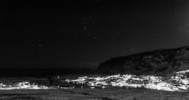 A nighttime scene of a small seaside village illuminated with artificial lights. The village is nestled at the base of a large, dark cliff, with a vast sky full of stars overhead. The landscape around the village appears to be rocky and rugged.
