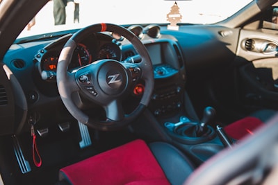 The interior of a sports car featuring a black steering wheel with the letter 'Z' logo in the center. The dashboard includes several gauges and a digital display. The car has a manual gear shift, and red accents on the seats are visible. Air freshener shaped like a tree hangs from the rearview mirror, and sunlight illuminates parts of the interior.
