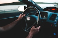 A person is gripping the steering wheel of a Honda car, with a view of the interior dashboard and partially visible speedometer. The scene outside the vehicle suggests a serene drive by a large body of water during dusk.
