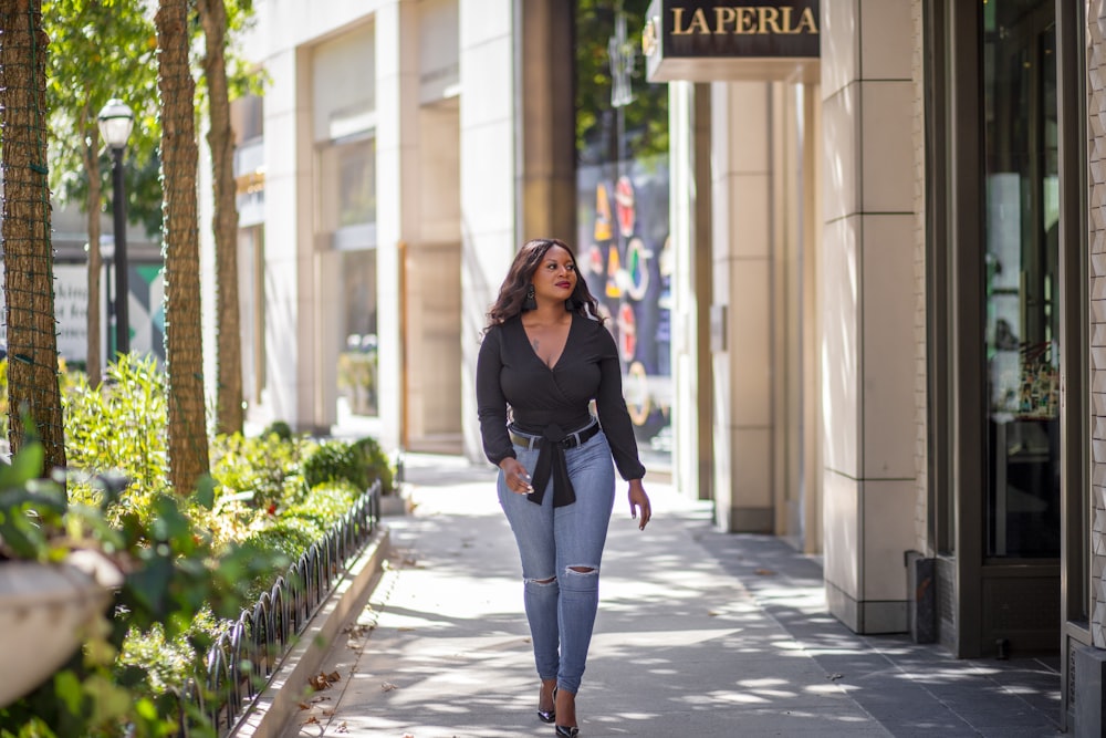 woman in black long sleeve shirt and blue denim jeans standing on sidewalk during daytime