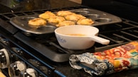 A muffin tray with baked goods, possibly muffins or cupcakes, is resting on a stove. Next to the tray is a white bowl with a spoon, filled with a yellow mixture. A colorful oven mitt with floral patterns lies nearby on the stove.