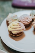 Several cupcakes are arranged on a white plate, each topped with swirls of pastel-colored frosting in shades of yellow, pink, and blue. The cupcakes appear to be vanilla or light brown, and the frosting is neatly piped to form elegant spiral patterns. The background is slightly out of focus, highlighting the pastel tones of the frosting against the soft surface of the plate.