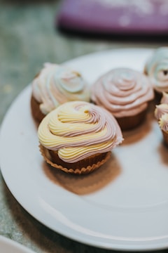 Several cupcakes are arranged on a white plate, each topped with swirls of pastel-colored frosting in shades of yellow, pink, and blue. The cupcakes appear to be vanilla or light brown, and the frosting is neatly piped to form elegant spiral patterns. The background is slightly out of focus, highlighting the pastel tones of the frosting against the soft surface of the plate.
