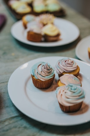 Three cupcakes with swirled pastel frosting are placed on a white plate. The cupcakes have a mix of blue, purple, and light pink frosting in a smooth, elegant design. In the background, additional cupcakes are slightly blurred, adding depth to the image with a hint of more colorful frosting.
