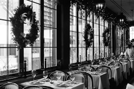 A monochromatic indoor scene of a restaurant with large windows decorated with festive wreaths. The dining tables are elegantly set with plates, glasses, and cutlery, and the chairs are neatly placed around the tables. A server is present on the right side of the image, seemingly setting up or attending to the tables.