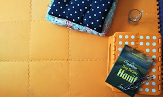 A soft, square polka-dotted cushion is placed on top of a floral-patterned textile laid out over a bright orange foam mat with interlocking edges. Nearby, a book with a green cover and text on the front, glasses, and a half-empty glass cup rest on an orange foldable stool.