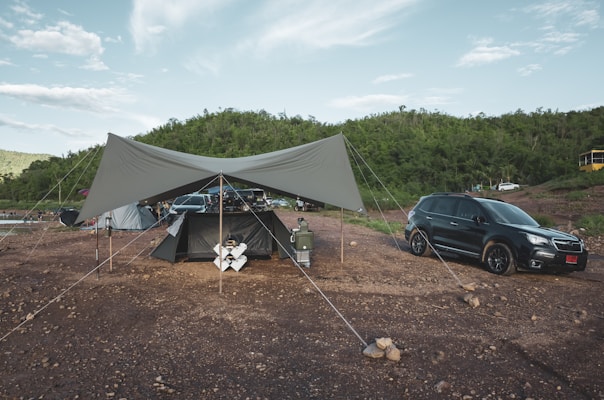 A camping setup with a large tarp canopy is erected on a rocky ground. Nearby, a black SUV is parked, and several camping gear items such as a cooler and chairs are visible. In the background, there is a lush green hillside under a partially cloudy sky.