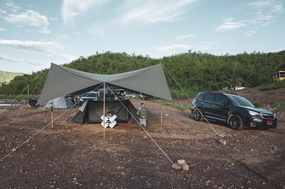 A camping setup with a large tarp canopy is erected on a rocky ground. Nearby, a black SUV is parked, and several camping gear items such as a cooler and chairs are visible. In the background, there is a lush green hillside under a partially cloudy sky.