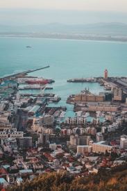 An aerial view of a coastal city with a busy harbor, featuring numerous buildings, docks, and ships. The sea extends toward the horizon, with a soft gradient of blues, and the city is dotted with various residential and commercial structures. The layout of roads and docks suggests a well-organized urban area with vibrant maritime activity.