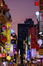 A bustling urban street filled with colorful neon signs in various languages, predominantly Korean. The signs advertise different businesses, from karaoke bars to restaurants. The street is lit with bright, vivid lights against the subtle hues of the evening sky. A mixture of commercial energy and cultural vibrancy is evident.