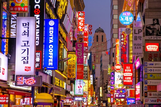 A bustling urban street filled with vibrant, colorful neon signs in Korean. The area is crowded with signs advertising various businesses like coffee shops, music bars, karaoke, and restaurants. The signs are stacked closely together, creating a lively and chaotic visual effect. The scene is set either in the evening or nighttime, as the signs are brightly lit against the dimming sky.