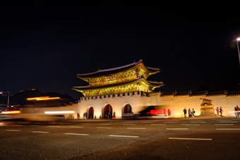 An illuminated traditional gate stands prominently against a dark night sky, with streaks of light from passing vehicles in front. The gate is elaborately decorated and brightly lit, drawing attention to its detailed architecture. People are visible near the gate, adding a sense of scale and activity.