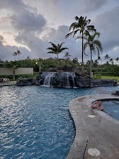 A swimming pool with a curved edge is set against a background of lush greenery and tall palm trees. In the pool, there is a waterfall feature cascading from rocks. A person is visible in the water near the rocks, and another person is standing at the edge of the pool near a hot tub or spa area. The sky is overcast with large, fluffy clouds.