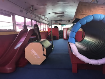An interior space of a bus converted into a play area with various playground equipment, including a slide and a tunnel. Soft padding and colorful designs make the environment child-friendly and safe for playtime.