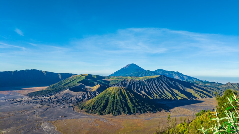 A stunning landscape featuring a volcanic mountain range under a clear blue sky. The main volcano exhibits a rugged, barren terrain mixed with patches of green vegetation at its base. Surrounding the foreground, the crater is detailed with patterned ridges, and sparse greenery can be seen along the edge.