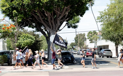 A group of people are walking along a street holding signs and banners, including one that reads 'No to Racism.' They appear to be participating in a peaceful protest or march. Trees line the street, and several cars are parked on the roadside. The atmosphere is bright and sunny, suggesting a daytime event. Several participants are wearing hats, and the group is diverse in age and attire.