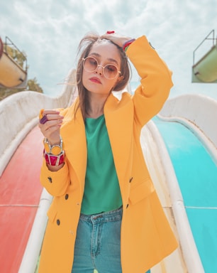 A fashionable woman wearing a bright yellow coat over a green top and blue jeans stands confidently with one hand in her hair. She wears stylish round sunglasses and large colorful bracelets, set against the backdrop of a playground slide with vibrant red and blue colors.
