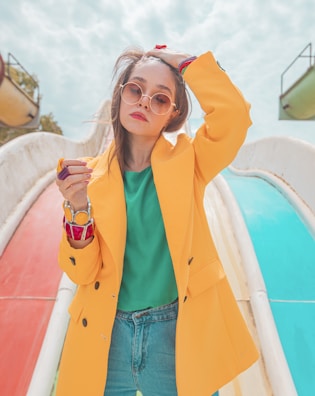 A fashionable woman wearing a bright yellow coat over a green top and blue jeans stands confidently with one hand in her hair. She wears stylish round sunglasses and large colorful bracelets, set against the backdrop of a playground slide with vibrant red and blue colors.